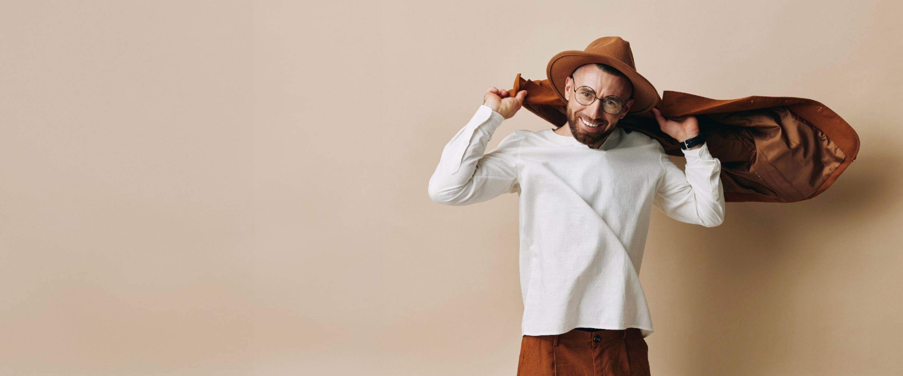 Smiling man wearing a white long-sleeve shirt, brown pants, and a stylish brown hat, exuding a modern casual look