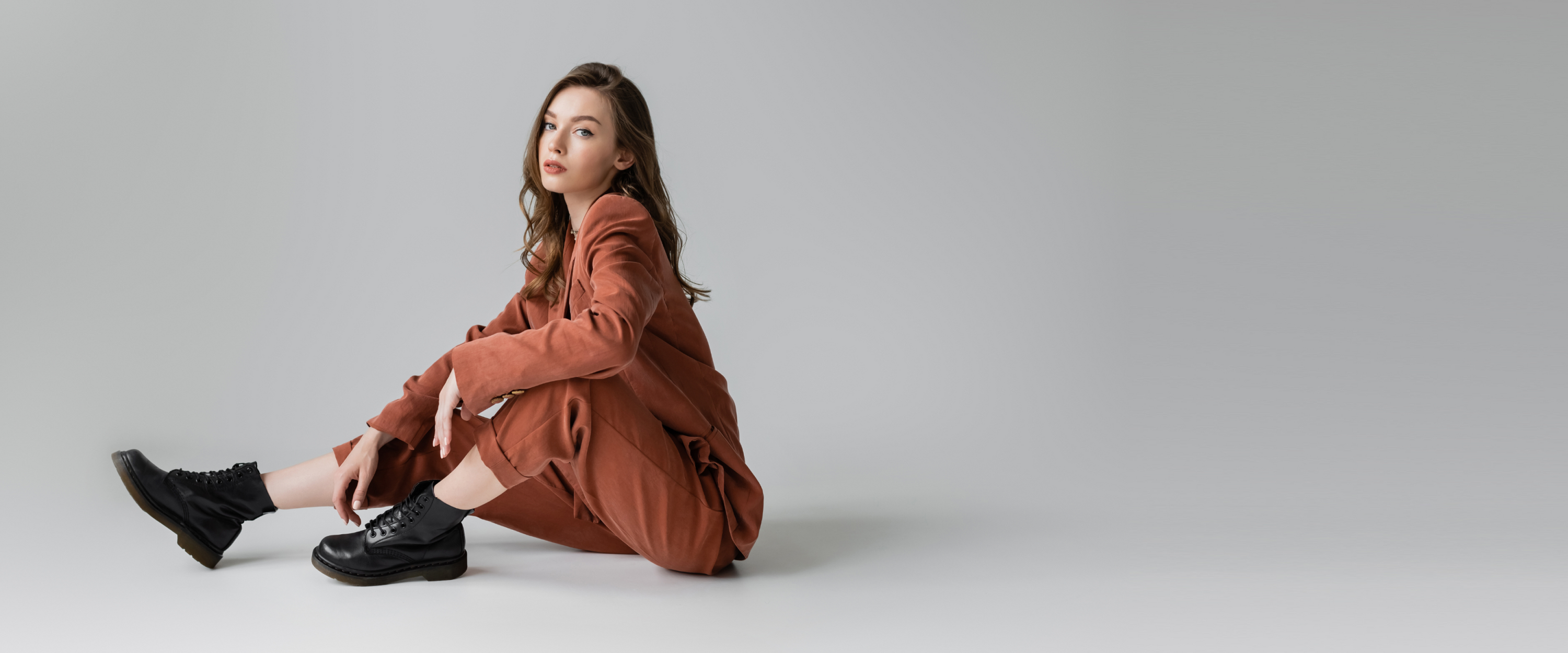 Young woman in a brown suit paired with black leather boots, sitting in a relaxed pose on a neutral background