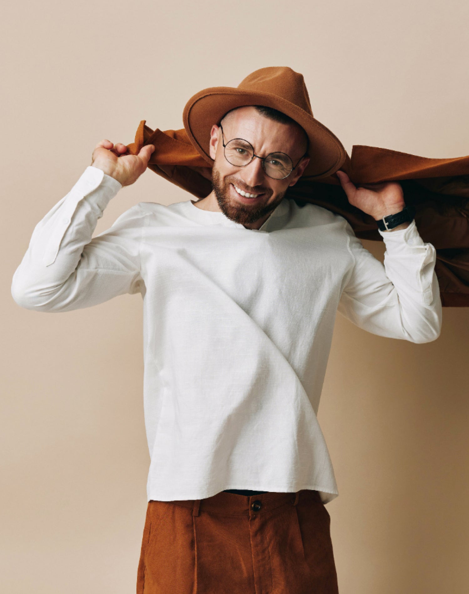 Smiling man wearing a white long-sleeve shirt, brown pants, and a stylish brown hat, exuding a modern casual look