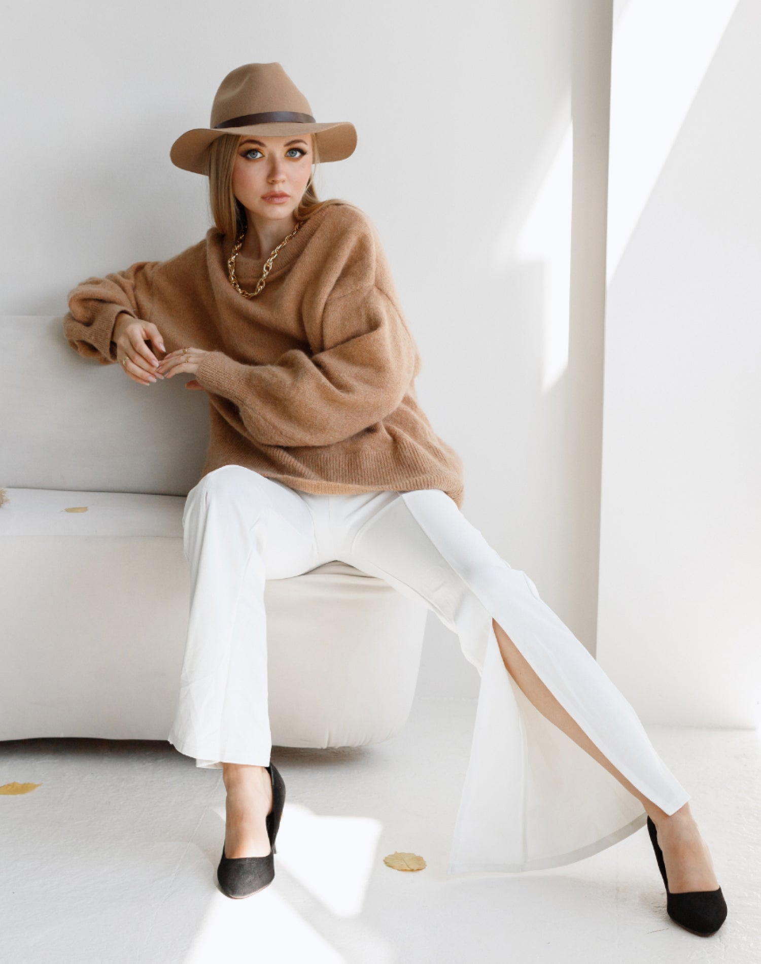 Elegant woman wearing a beige sweater, wide-brim hat, and white slit pants, accessorized with a gold chain necklace