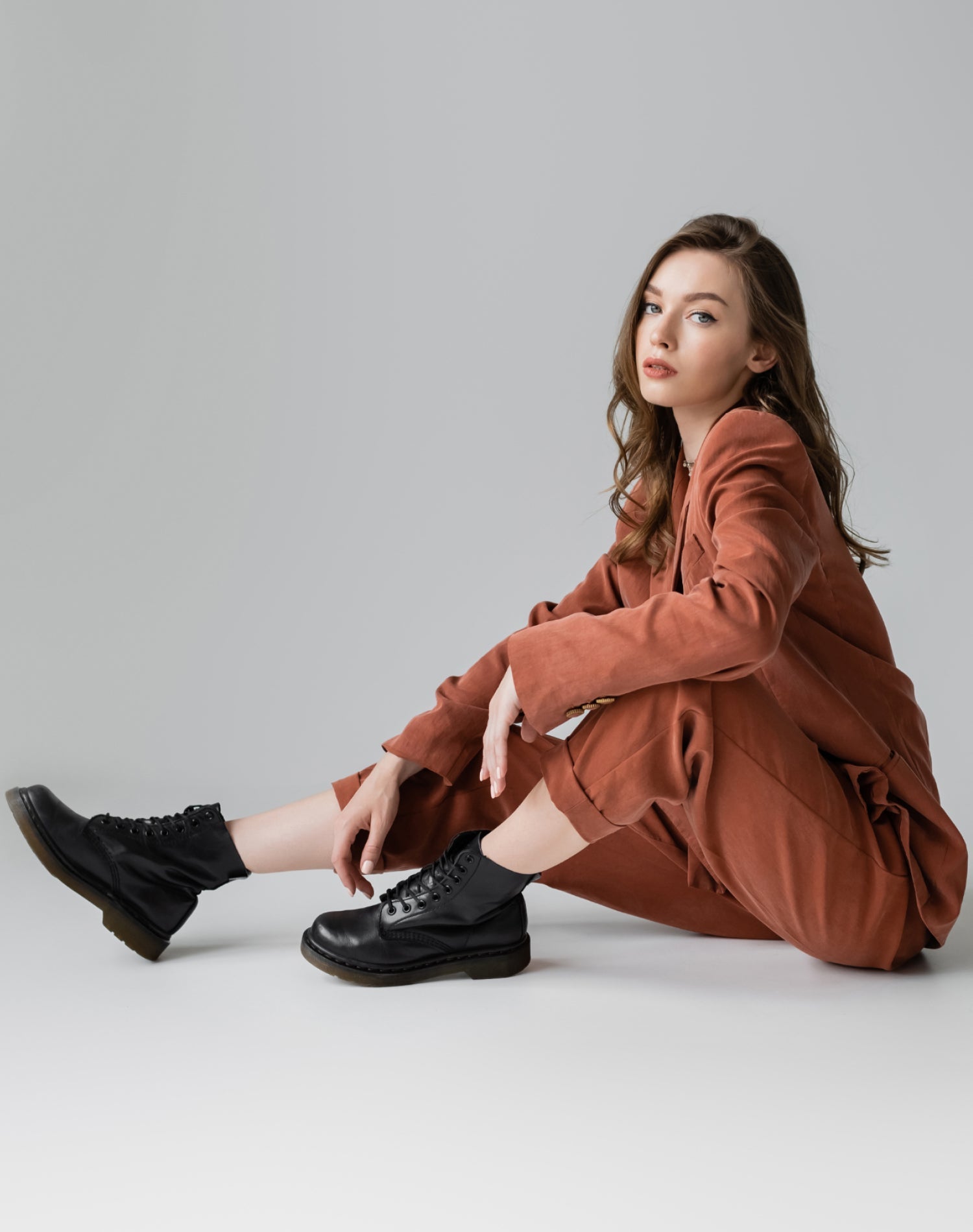 Young woman in a brown suit paired with black leather boots, sitting in a relaxed pose on a neutral background